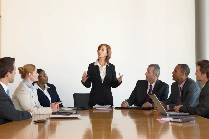 A woman standing at the head of a table with several other people.