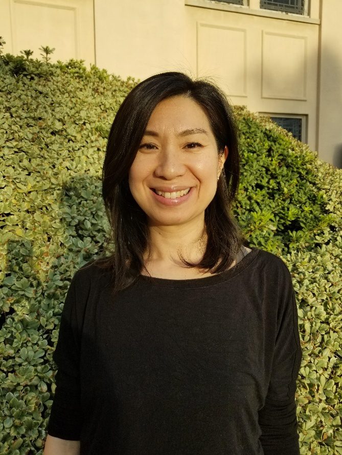 A woman standing in front of bushes smiling for the camera.